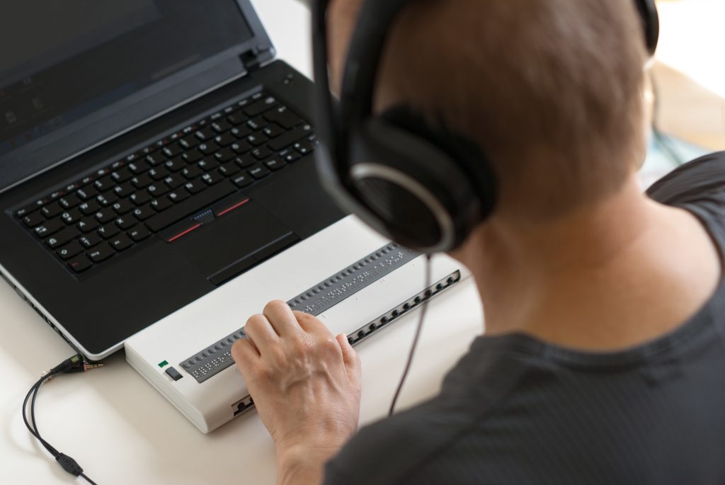 Over-the-shoulder view of a person wearing headphones and using a Braille display device connected to a laptop.