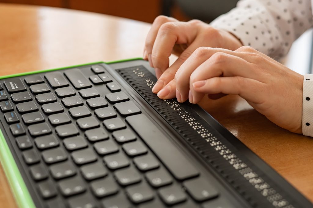 Close-up of a woman's hands reading a line of Braille on an electronic Braille display device integrated into a keyboard.
