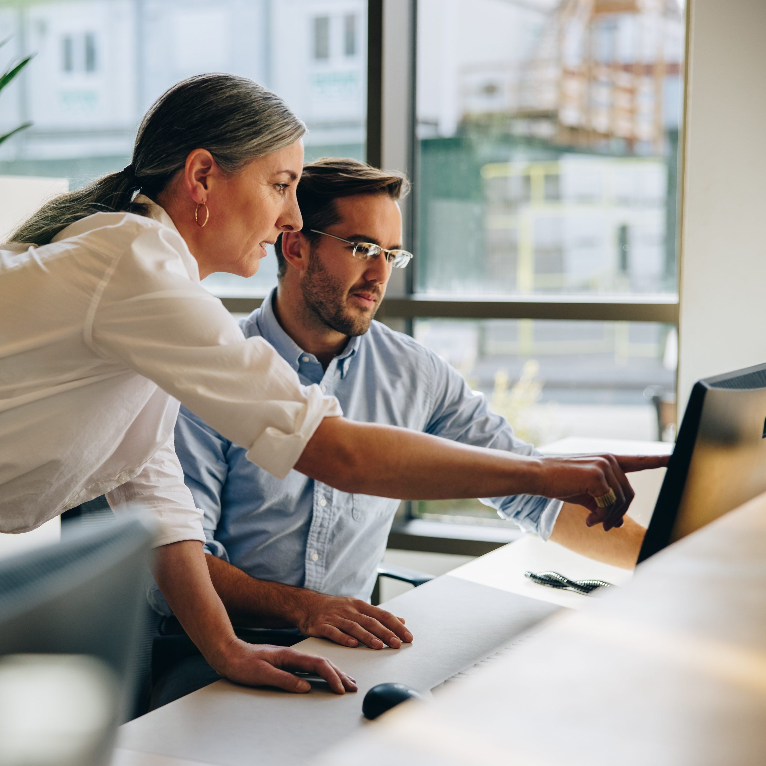 Woman pointing at a desktop monitor helping a colleague sitting at a desk.