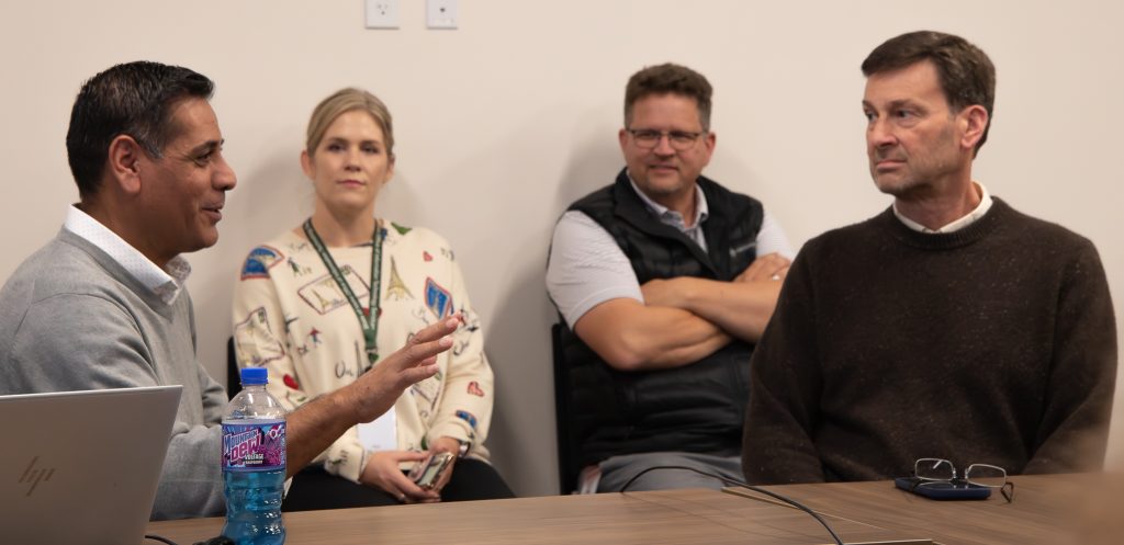 Four adults in the meeting engage in a focused discussion around a table with a laptop and drink.