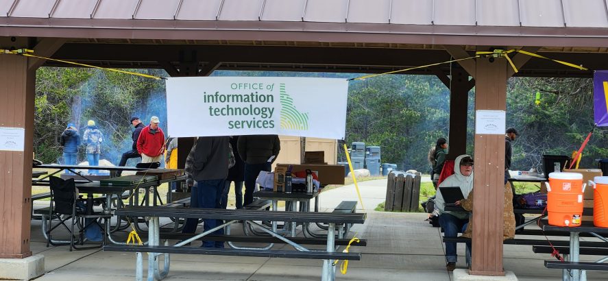 Outdoor pavilion with an Information Technology Services banner, picnic tables, people gathered, equipment and coolers set up, and trees in the background.