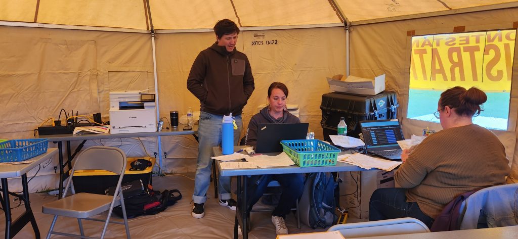 People working inside a tent with laptops, paperwork, and office equipment on tables, creating a temporary workspace setup.