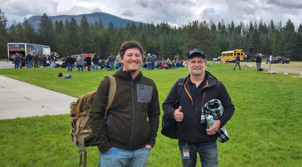 Two people standing on a grassy field with backpacks and gear, with a large group, vehicles, and forested mountains in the background.