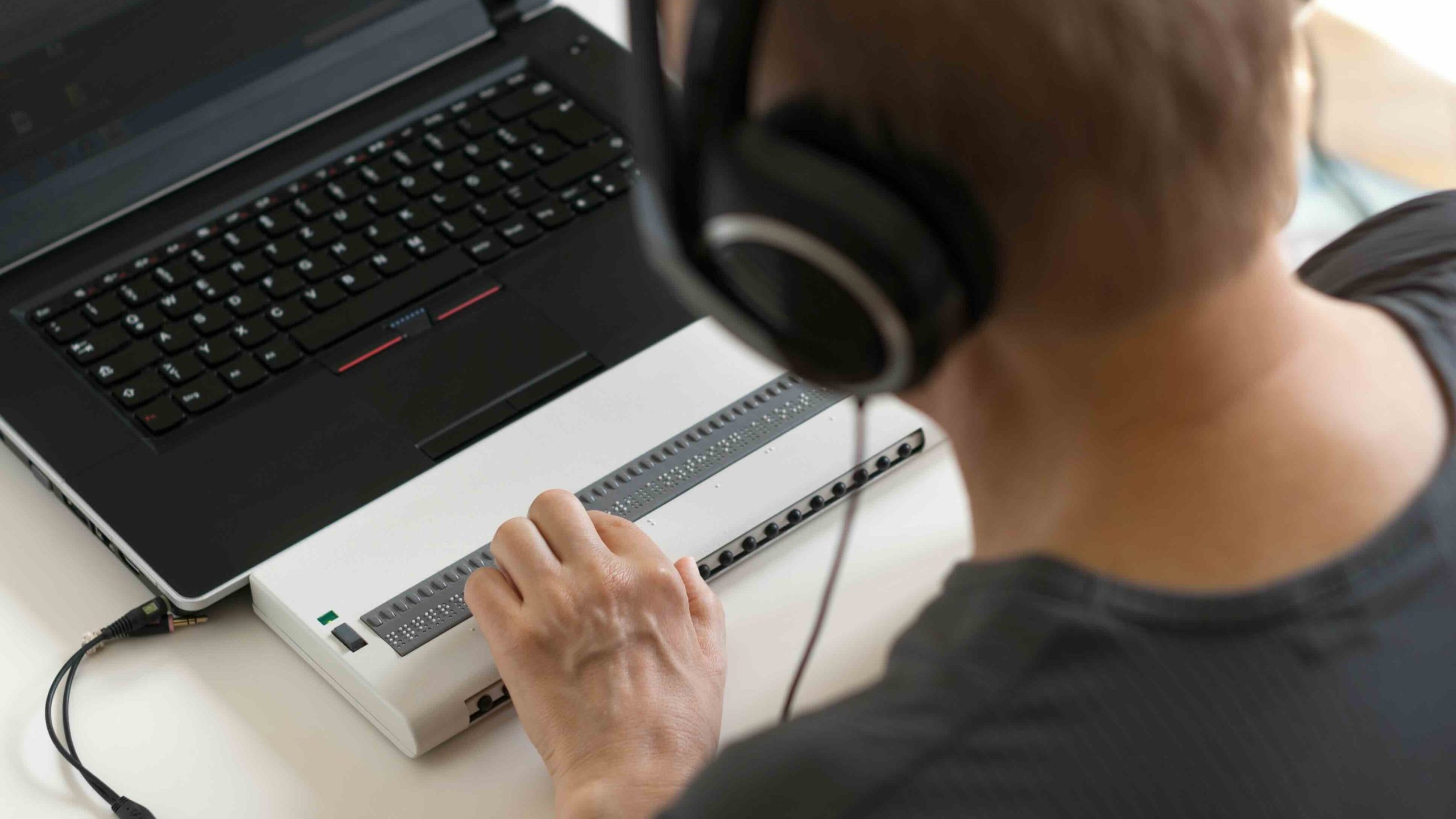 Person using a laptop with a refreshable braille display and wearing large over-ear headphones, with one hand reading the braille device on a desk.