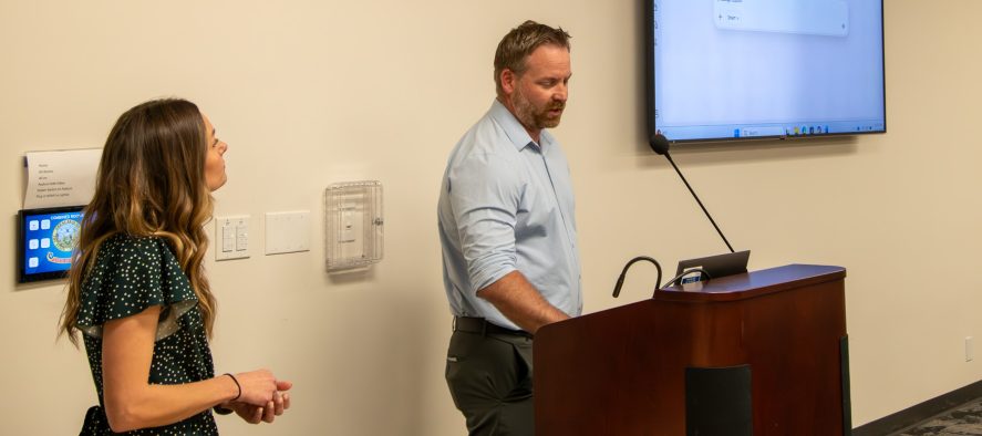 Taylor Bothke abd James Palmer stand at the front of a room near a podium and large screen giving a presentation.